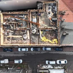 Zdjęcie z lotu ptaka przedstawia plac budowy z widocznymi fundamentami i szalunkami, zbrojeniami oraz elementami drewnianymi przygotowanymi do zalewania betonem. Na ulicy zaparkowane samochody.