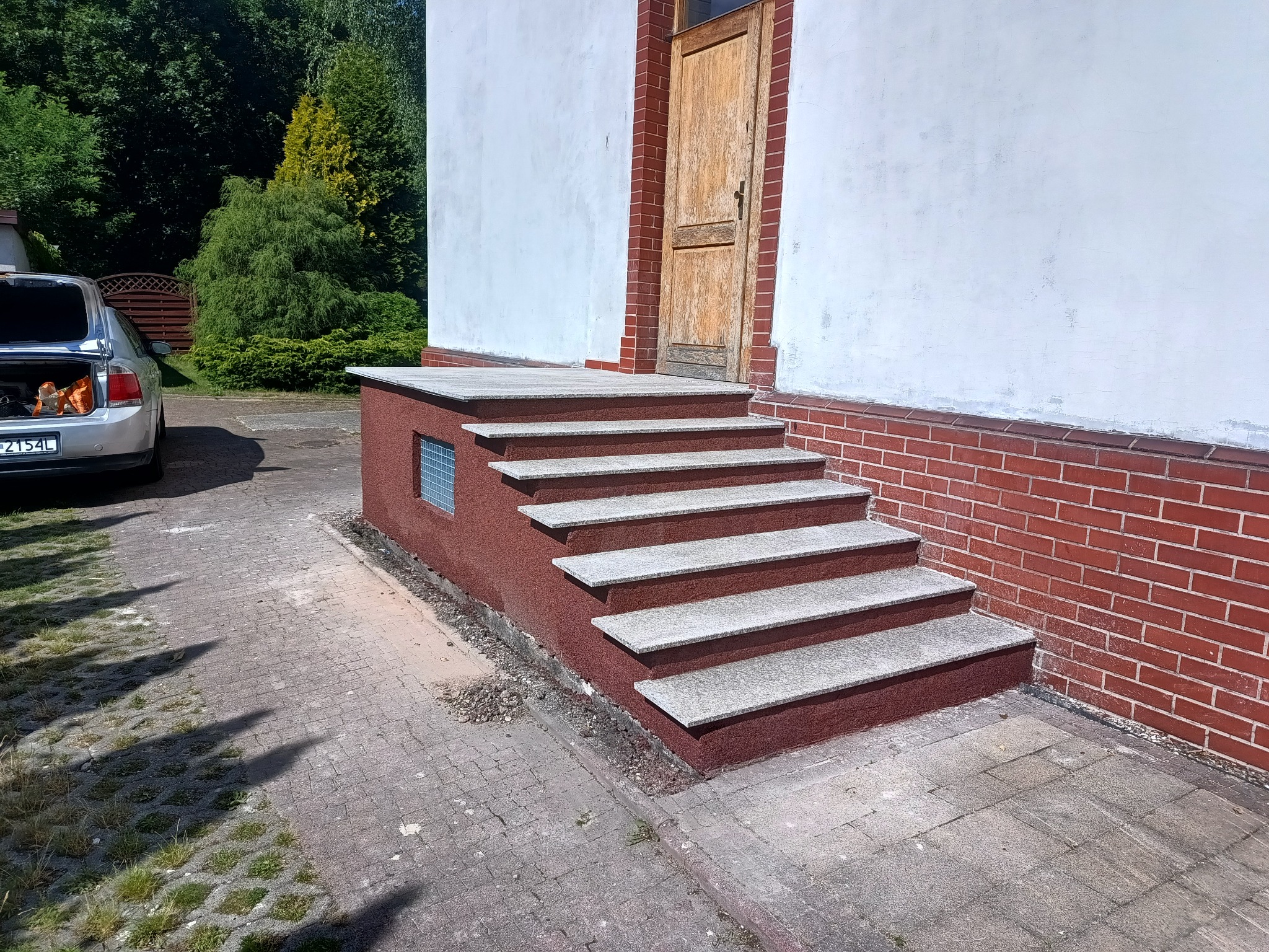 Zewnętrzne schody z szarego kamienia prowadzące do drewnianych drzwi, z boku ściana z czerwonej cegły i srebrny samochód w tle.