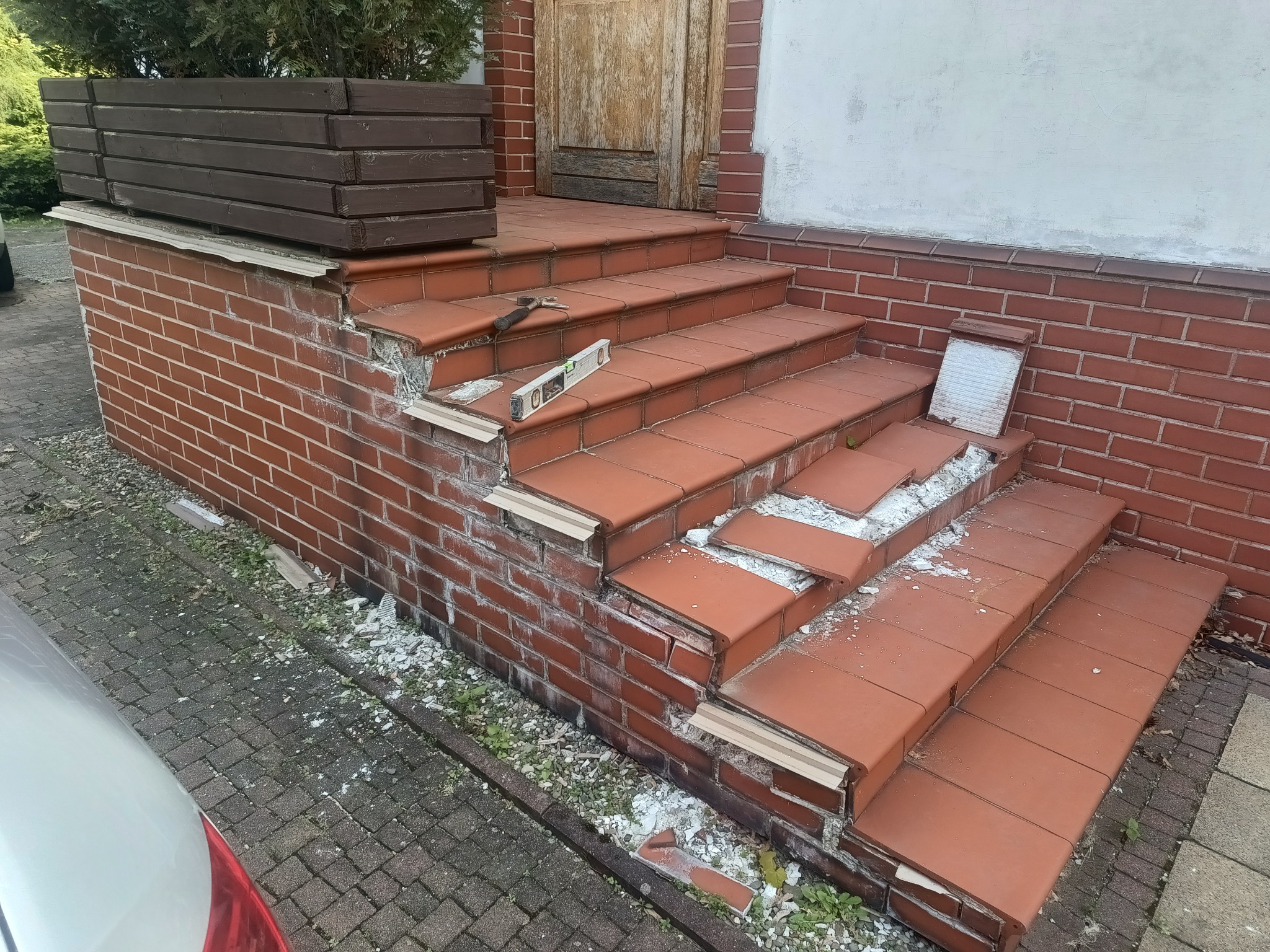 Uszkodzone, ceglane schody wejściowe z poziomnicą i młotkiem na jednym ze stopni, fragmenty płytek porozrzucane wokół podstawy.