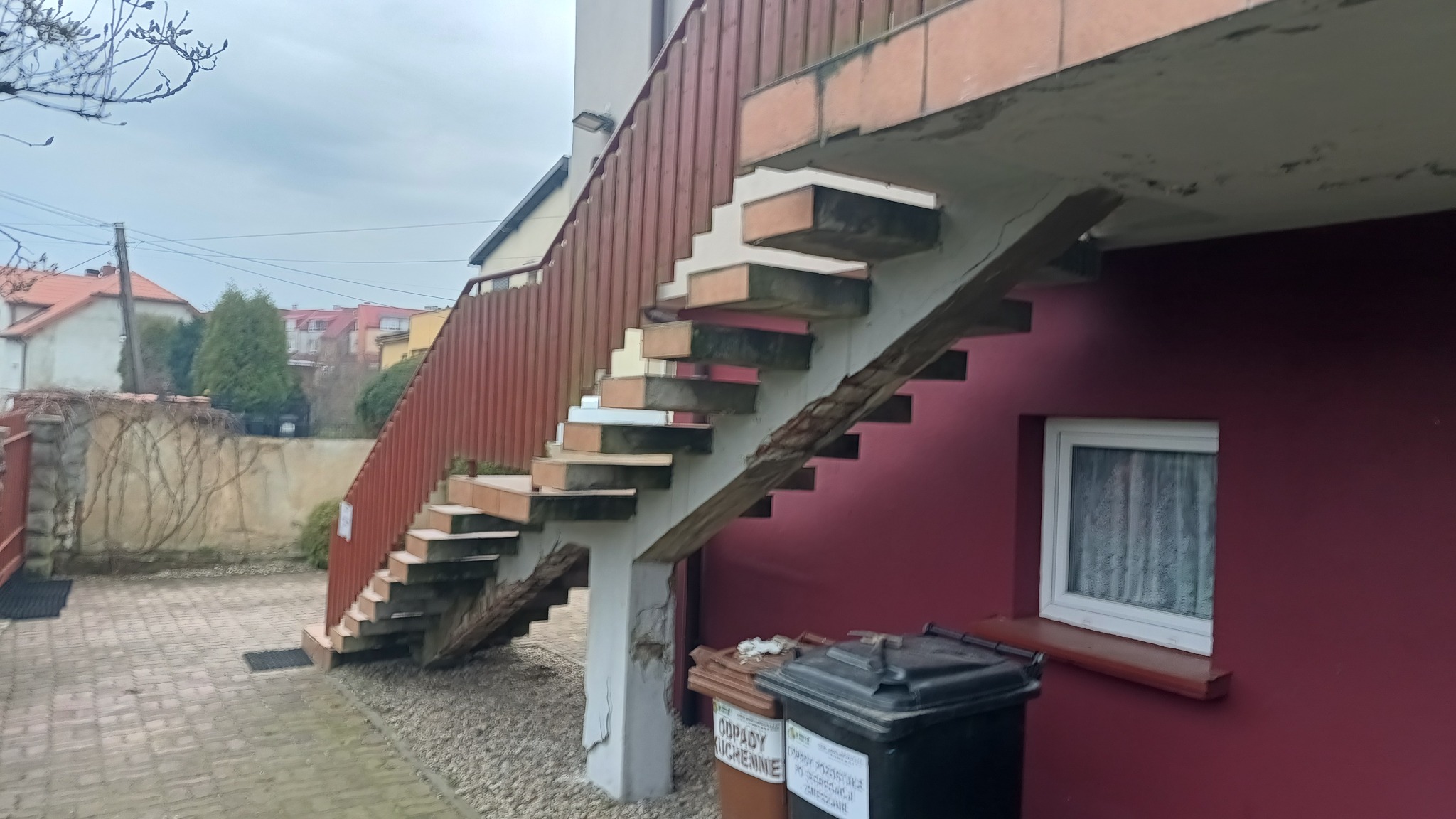 Zewnętrzne schody z płytkami w trakcie remontu, widoczne uszkodzone elementy konstrukcyjne i przygotowane stopnie, obok ściana budynku z oknem i kosze na śmieci.
