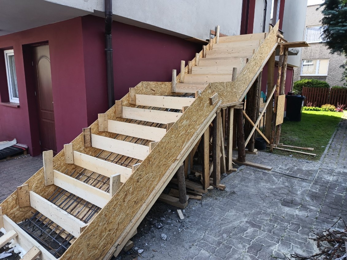 Przygotowanie do wylania betonowych schodów zewnętrznych: szalunek z płyt OSB, drewniane podpory i widoczna siatka zbrojeniowa wewnątrz formy.