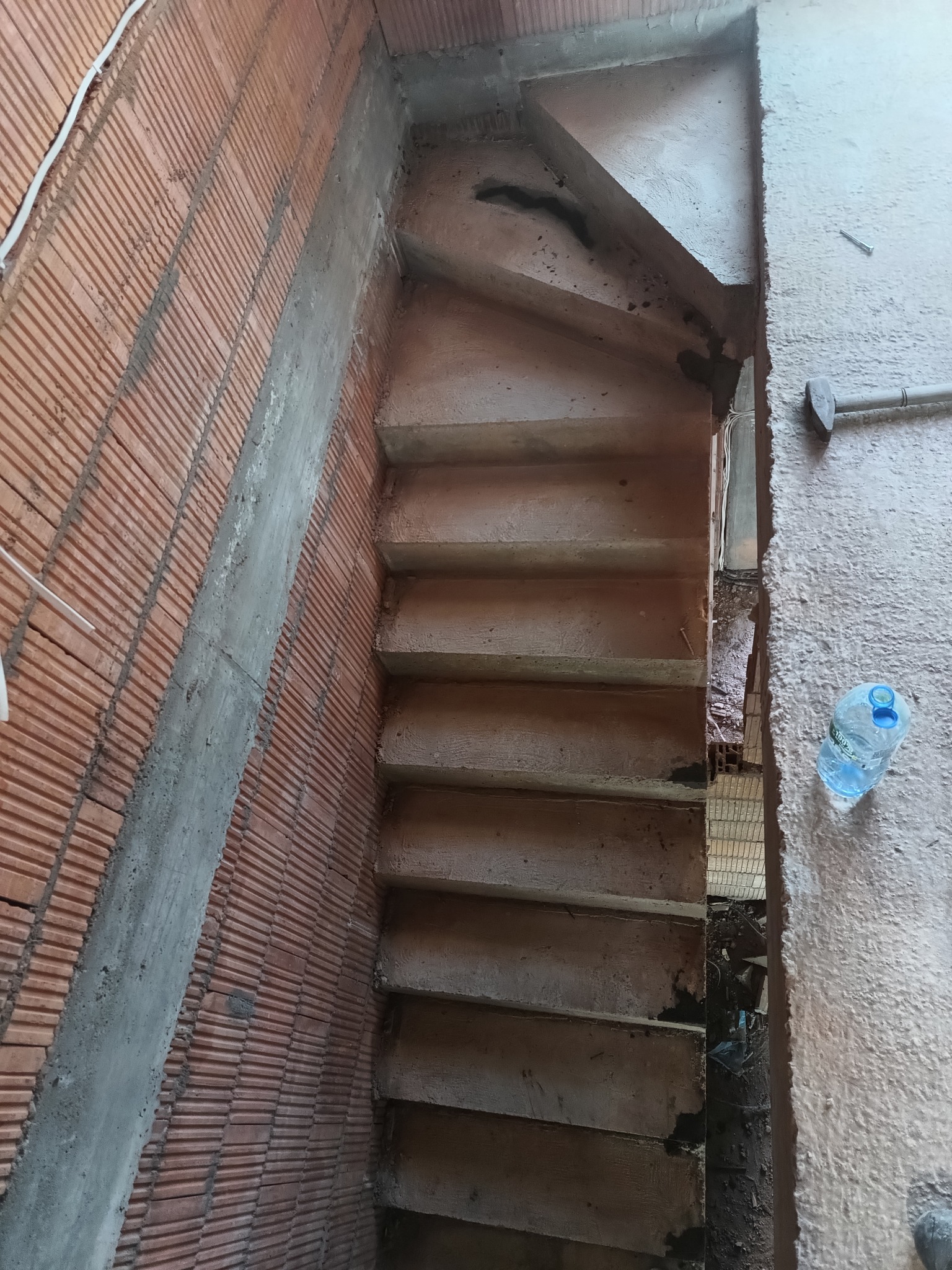 Wewnętrzne schody betonowe w surowym stanie, widoczne ślady budowy, ściany z cegły dziurawki i butelka wody obok.