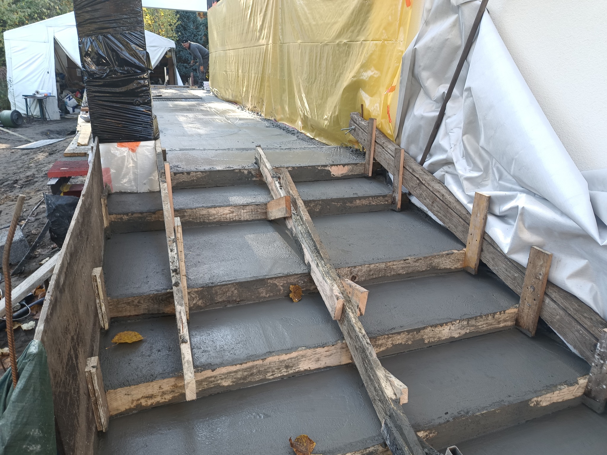 Wylewanie betonu w drewnianym szalunku na schodach zewnętrznych, widoczne liście na świeżym betonie, w tle folia ochronna i namiot budowlany.
