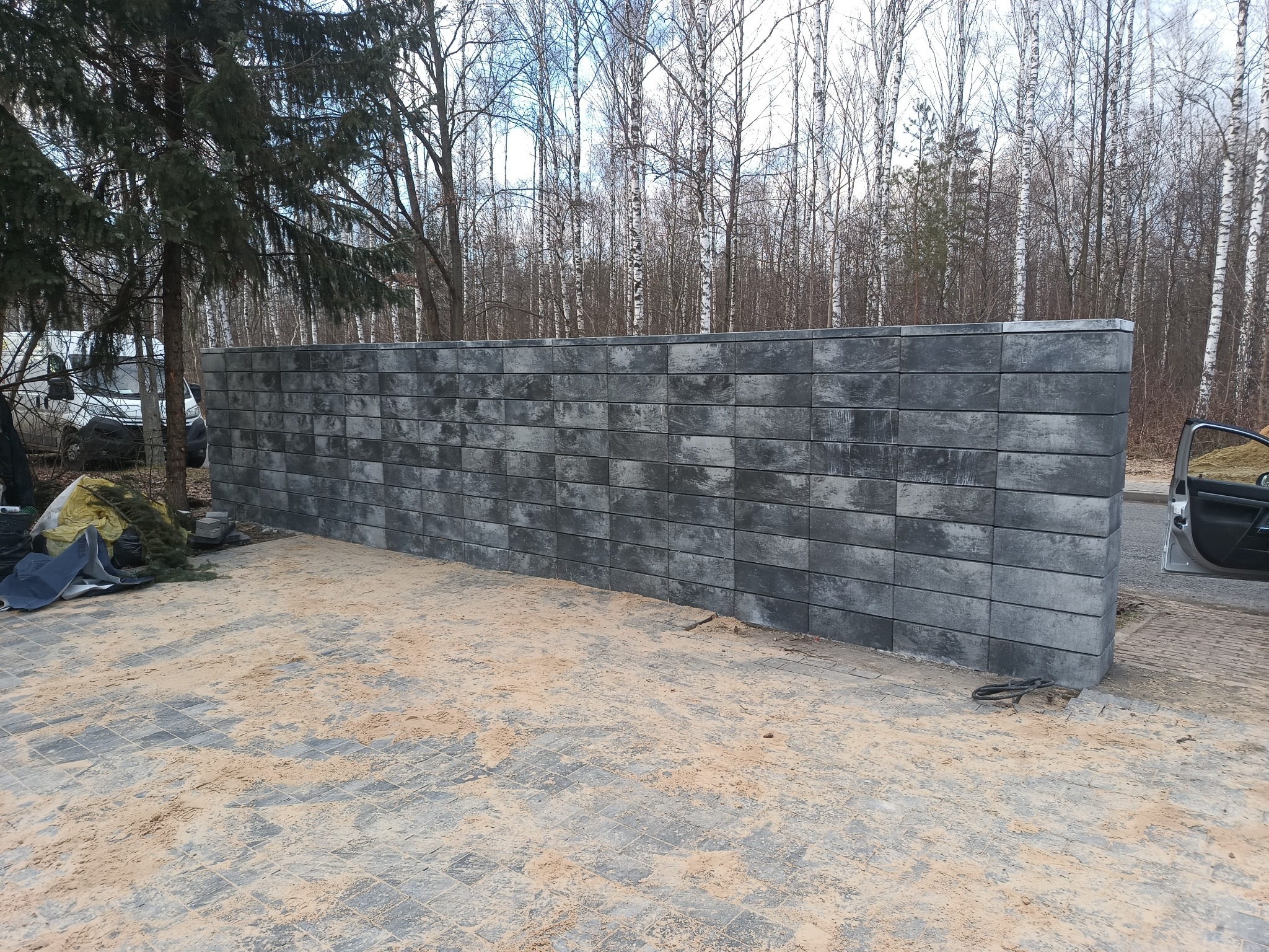 Nowo wybudowany, szary mur oporowy z betonowych bloczków, widoczny na tle drzew i częściowo zaparkowanego samochodu. Podłoże wyłożone kostką brukową pokryte piaskiem.