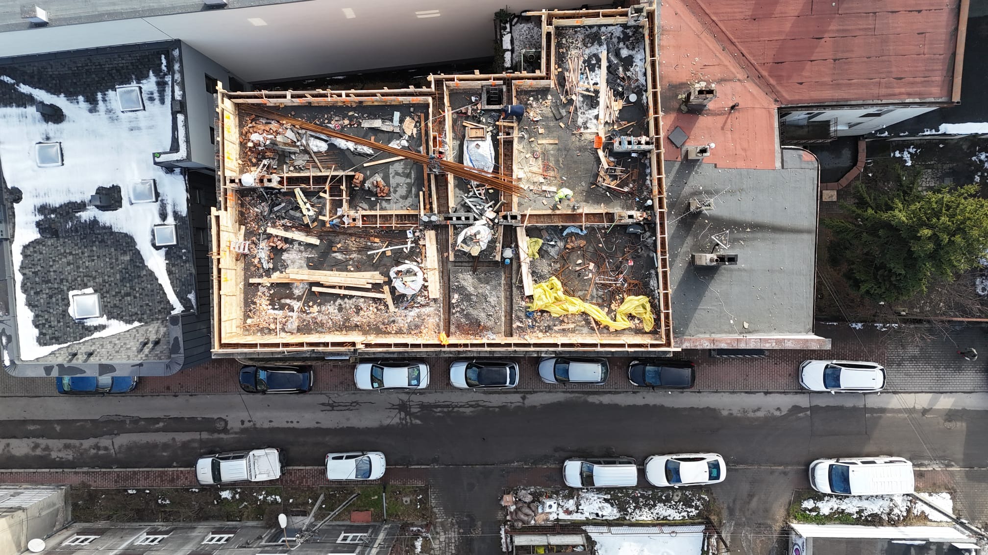 Zdjęcie z lotu ptaka przedstawia plac budowy z widocznymi fundamentami i szalunkami, zbrojeniami oraz elementami drewnianymi przygotowanymi do zalewania betonem. Na ulicy zaparkowane samochody.