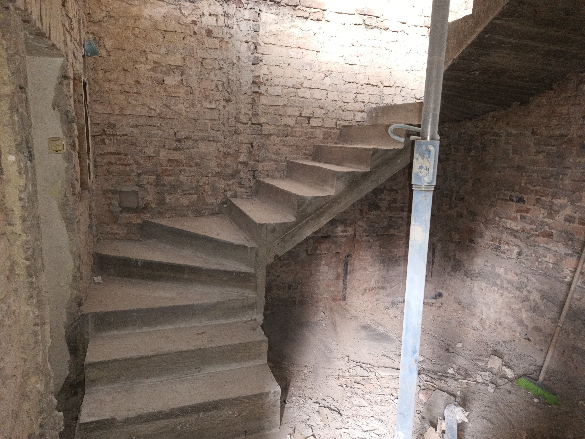 Wewnętrzne spiralne schody z surowego betonu w trakcie budowy, wsparte metalową podporą, z widocznymi ceglanymi ścianami w stanie surowym i pozostałościami gruzu.