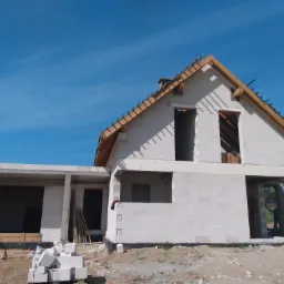 Surowy stan domu z białych bloczków na etapie budowy, widoczne drewniane elementy konstrukcji dachu i niezabezpieczone otwory okienne na tle błękitnego nieba.