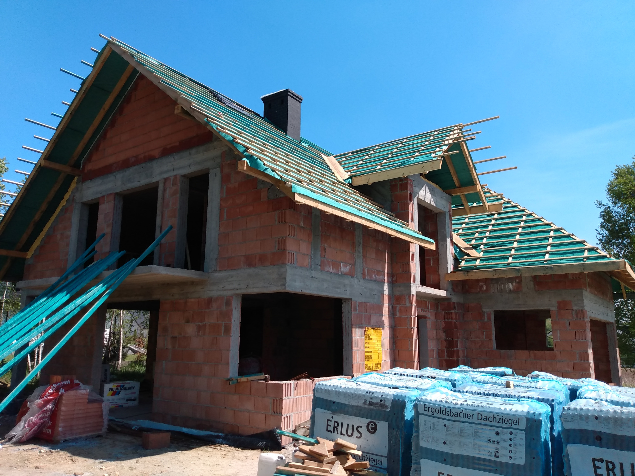 Surowy stan budowy domu z czerwonej cegły z widoczną konstrukcją dachu pokrytą zieloną membraną, paletami dachówek Erlus i stosem niebieskich rur w tle.