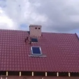 Czerwony dach z dachówki ceramicznej z dwoma oknami dachowymi i kominem z cegły, widoczny fragment ściany budynku z listwami przygotowanymi pod elewację, drabina oparta o ścianę.