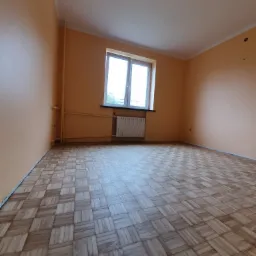 Pokój z drewnianą parkietową podłogą ułożoną w kwadraty, pomarańczowe ściany, białe okno i grzejnik. Widoczne rury grzewcze przy ścianie.