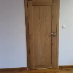 Zamknięte, dębowe drzwi wewnętrzne z prostokątną klamką, osadzone w białej ścianie nad drewnianą podłogą z widocznymi gwoździami.