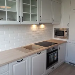 Klasyczna biała kuchnia z drewnianym blatem, białymi kaflami na ścianie, szklanymi frontami szafek wiszących i wbudowanym piekarnikiem oraz mikrofalówką.