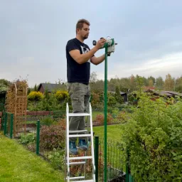 Instalator na drabinie w Warszawie montuje białą kamerę na zielonym słupku w ogrodzie, widoczne narzędzia i otoczenie domu.
