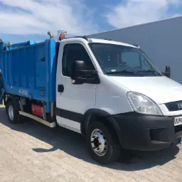 Biała śmieciarka Iveco z niebieską zabudową typu Eko-Cel, zaparkowana na kostce brukowej w słoneczny dzień, widoczny napis 'Odpady' na tablicy z przodu pojazdu.