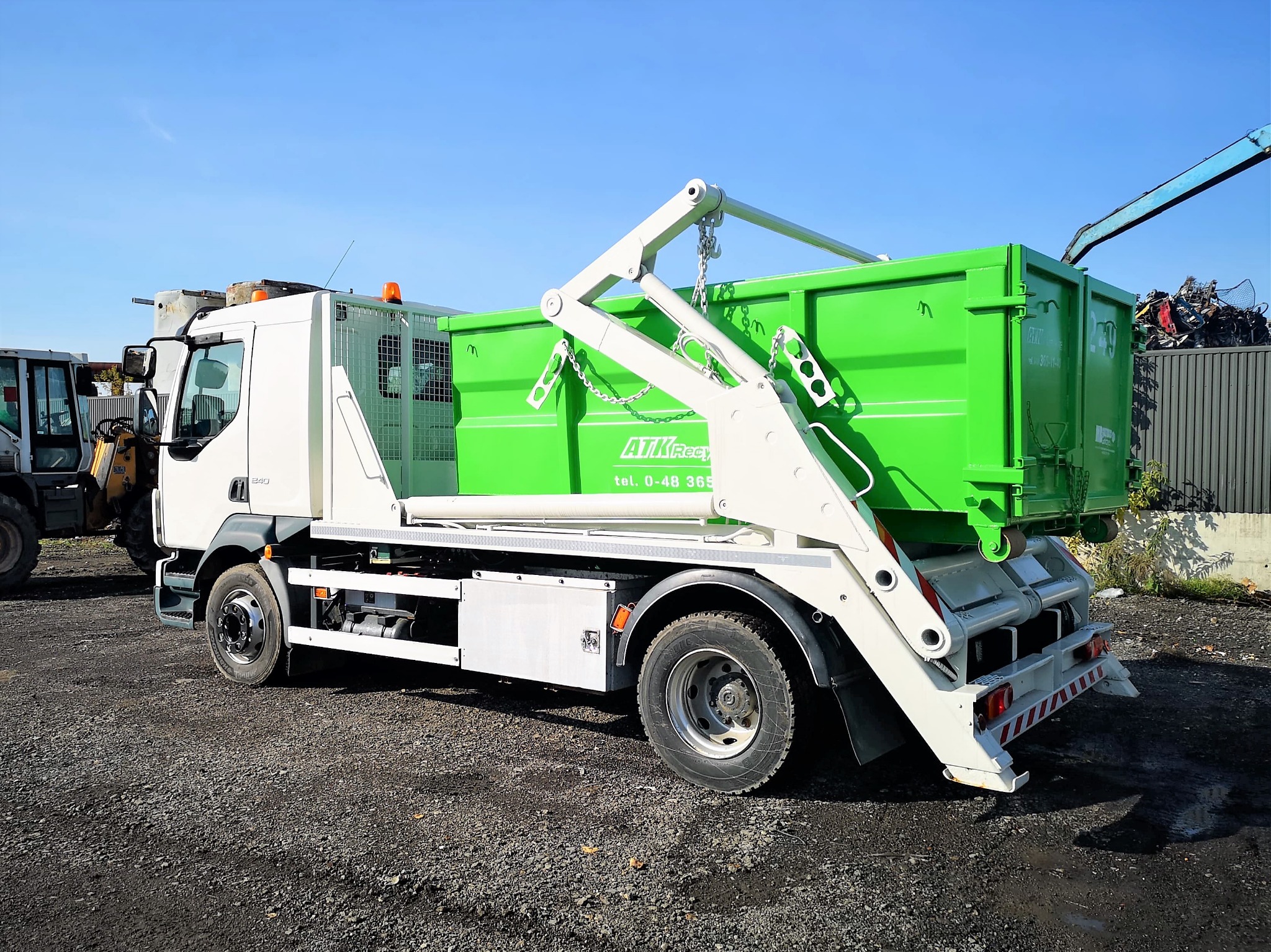 Biała ciężarówka z zielonym kontenerem na gruz, widok z boku na żwirowym terenie, w tle składowisko metalu i błękitne niebo.