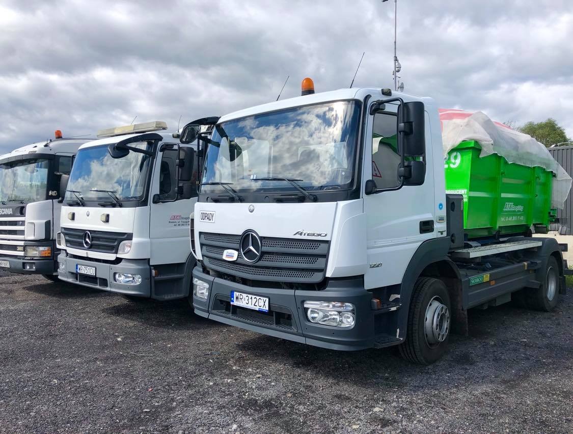 Trzy ciężarówki: Scania i dwa Mercedesy Atego, z czego jeden z zielonym kontenerem zabezpieczonym białą plandeką, stojące na żwirowym placu pod pochmurnym niebem.