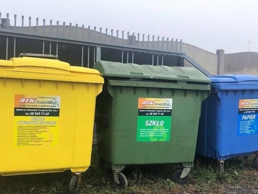 Trzy kolorowe kontenery na odpady segregowane: żółty na metale i tworzywa sztuczne, zielony na szkło i niebieski na papier, ustawione obok siebie na zewnątrz, z logo firmy ATK Recykling na każdym...
