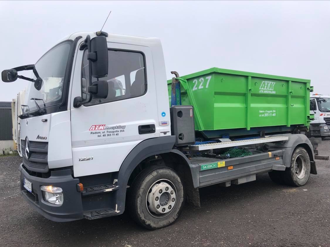 Biała ciężarówka Mercedes Atego z zielonym kontenerem oznaczonym numerem 227, należąca do firmy ATK Recykling z Radomia, stojąca na placu.