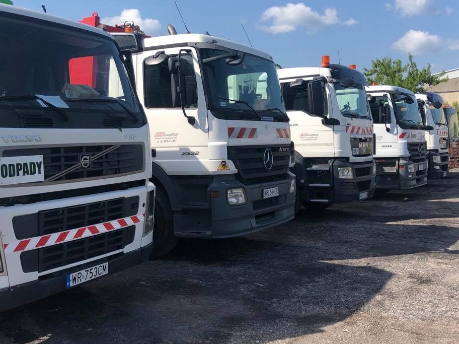 Szereg białych ciężarówek różnych marek (Volvo, Mercedes, MAN) z oznaczeniami firmowymi i ostrzegawczymi, zaparkowanych na placu pod błękitnym niebem.