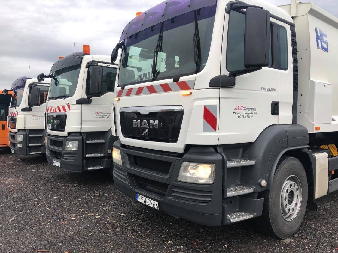 Rząd białych ciężarówek MAN z pomarańczowymi kogutami i oznaczeniami odblaskowymi, zaparkowanych na placu. Na kabinach widoczne logo firmy ATM Recycling z Radomia.