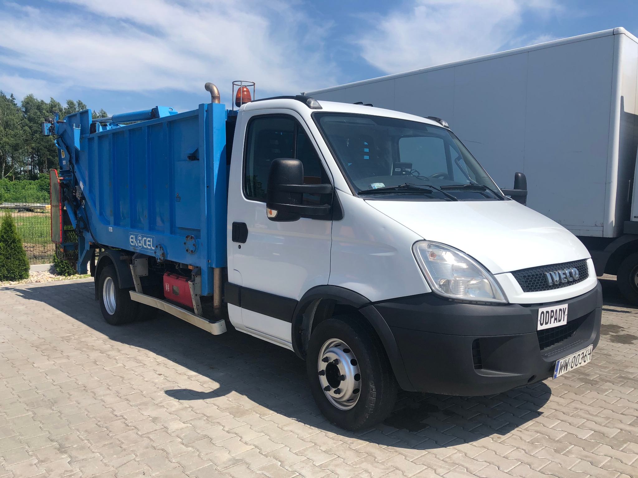 Biała śmieciarka Iveco z niebieską zabudową typu Eko-Cel, zaparkowana na kostce brukowej w słoneczny dzień, widoczny napis 'Odpady' na tablicy z przodu pojazdu.