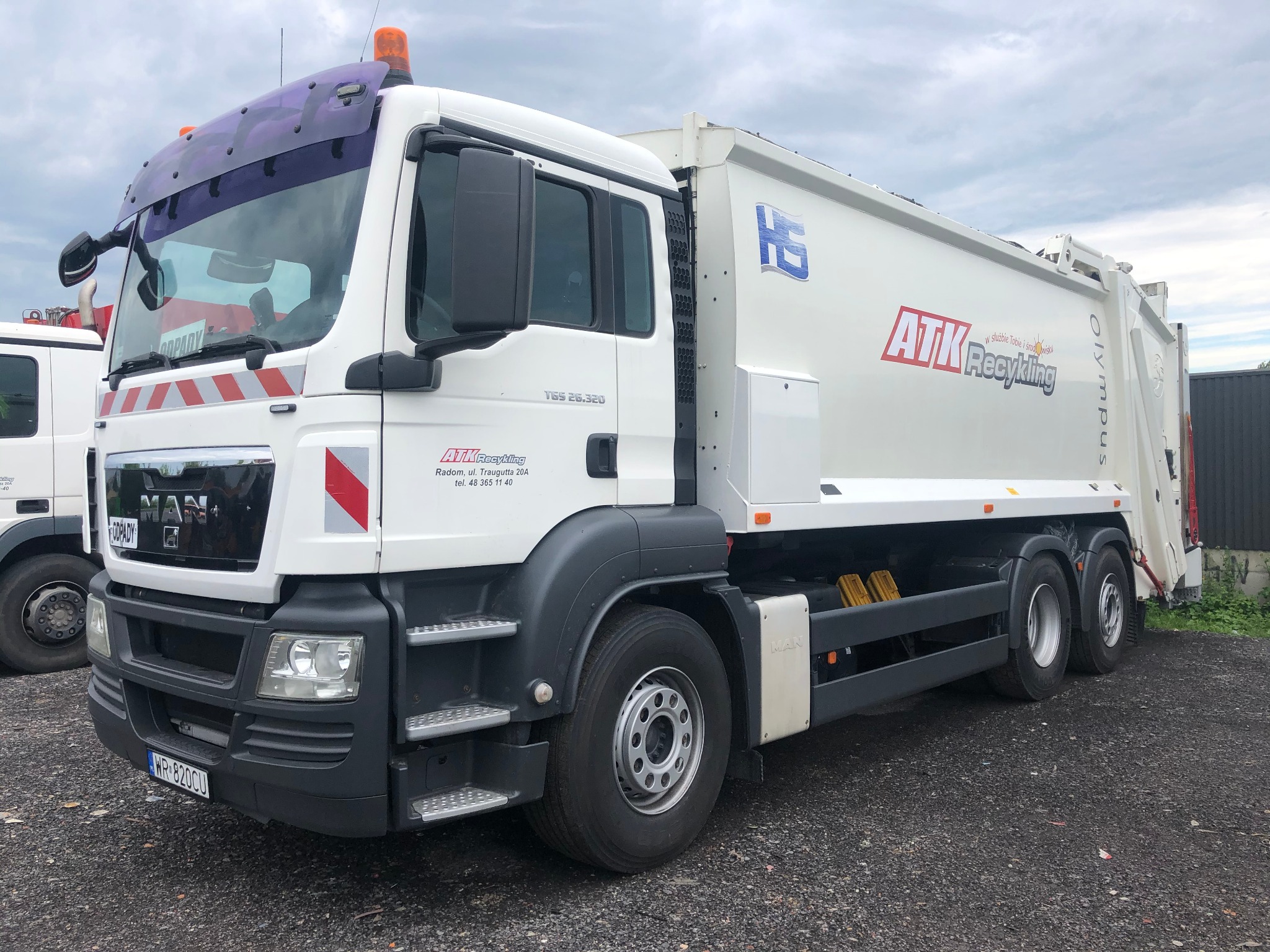 Biała śmieciarka MAN z logo ATK Recykling zaparkowana na żwirowym placu, widok z boku na przód pojazdu, z pomarańczowym światłem ostrzegawczym na dachu kabiny.