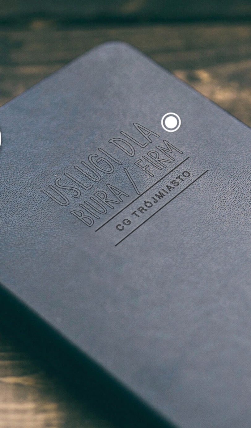 Ciemnoszary notes z wytłoczonym napisem 'Usługi dla biura / firm CG Trójmiasto' leżący na drewnianym blacie.