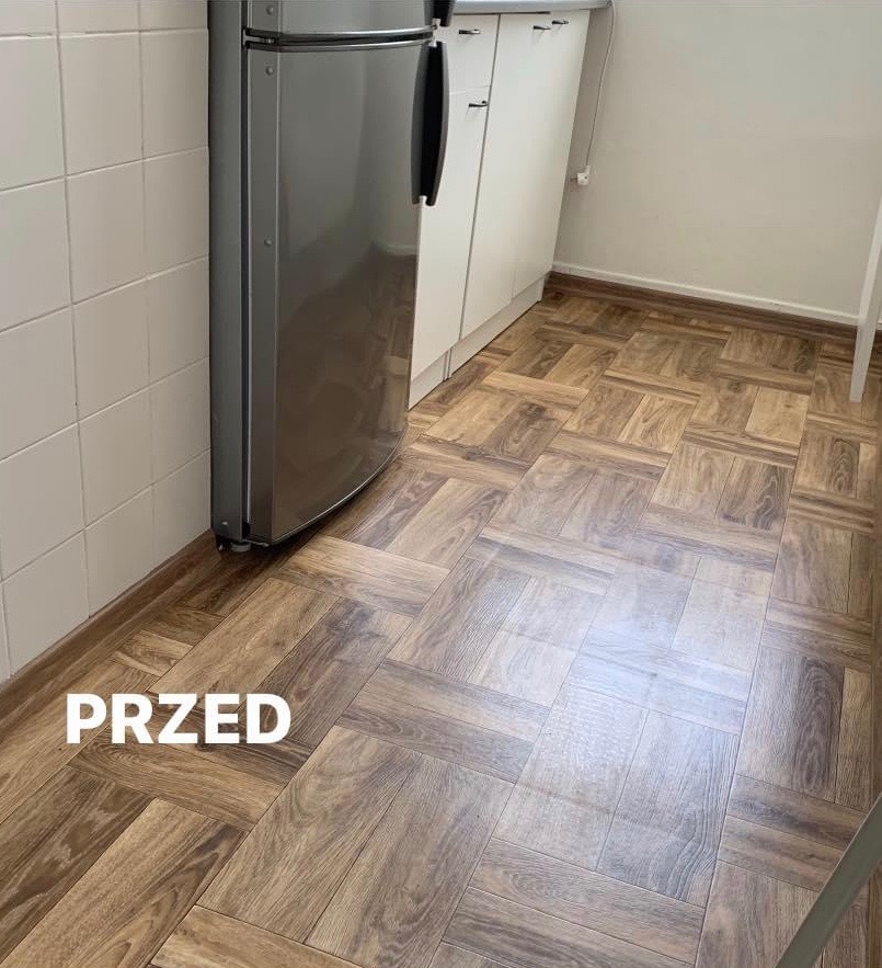 Kuchnia przed: brązowa podłoga imitująca drewno ułożona w kwadraty, białe płytki na ścianie, srebrna lodówka i białe szafki kuchenne.
