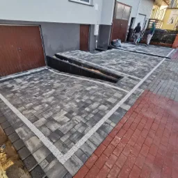 Nowo ułożona, szara kostka brukowa przed budynkiem mieszkalnym z liniowymi odwodnieniami i rampą wjazdową, obramowana białym grysem i przechodząca w czerwoną kostkę, z widocznymi pracownikami w tle.