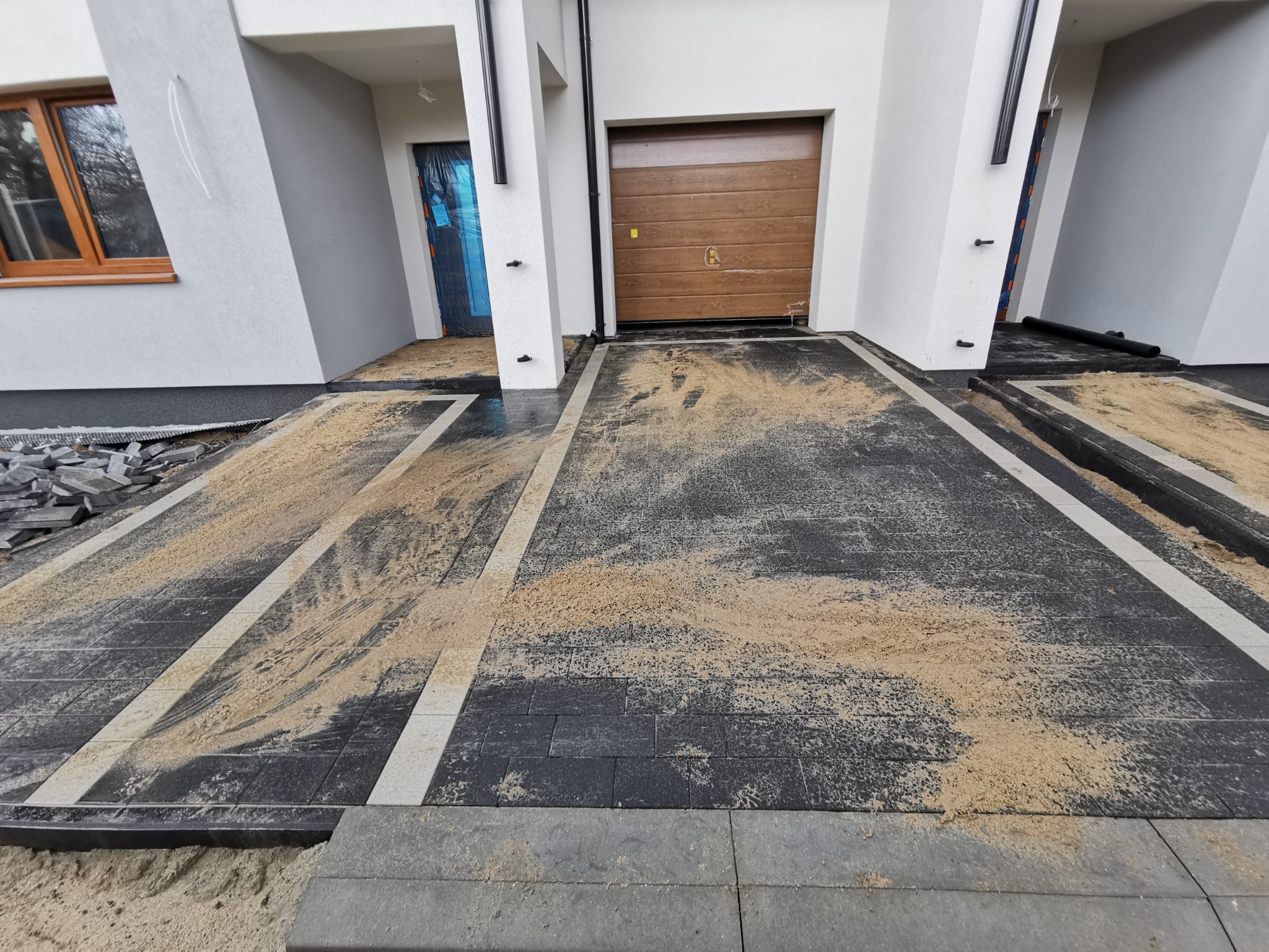 Nowoczesny podjazd z ciemnej kostki brukowej z jasnymi pasami, częściowo zasypany piaskiem, przed garażem i wejściem do domu w trakcie prac wykończeniowych.