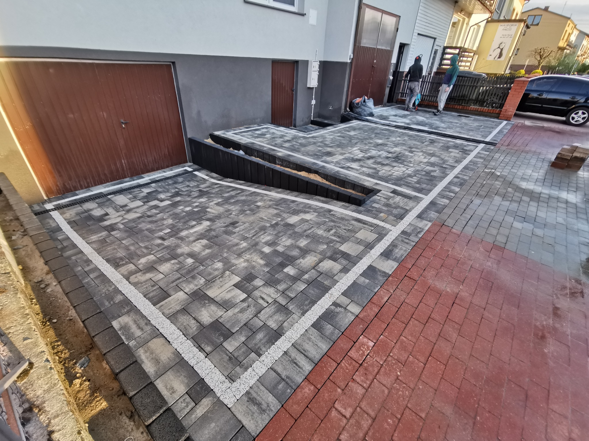 Nowo ułożona, szara kostka brukowa przed budynkiem mieszkalnym z liniowymi odwodnieniami i rampą wjazdową, obramowana białym grysem i przechodząca w czerwoną kostkę, z widocznymi pracownikami w tle.