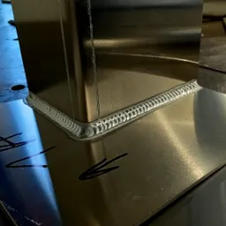 Połączenie spawane dwóch elementów z blachy nierdzewnej pod kątem prostym, widoczny regularny ścieg spawalniczy typu TIG, na jednej z blach widoczne odręczne oznaczenia markerem.