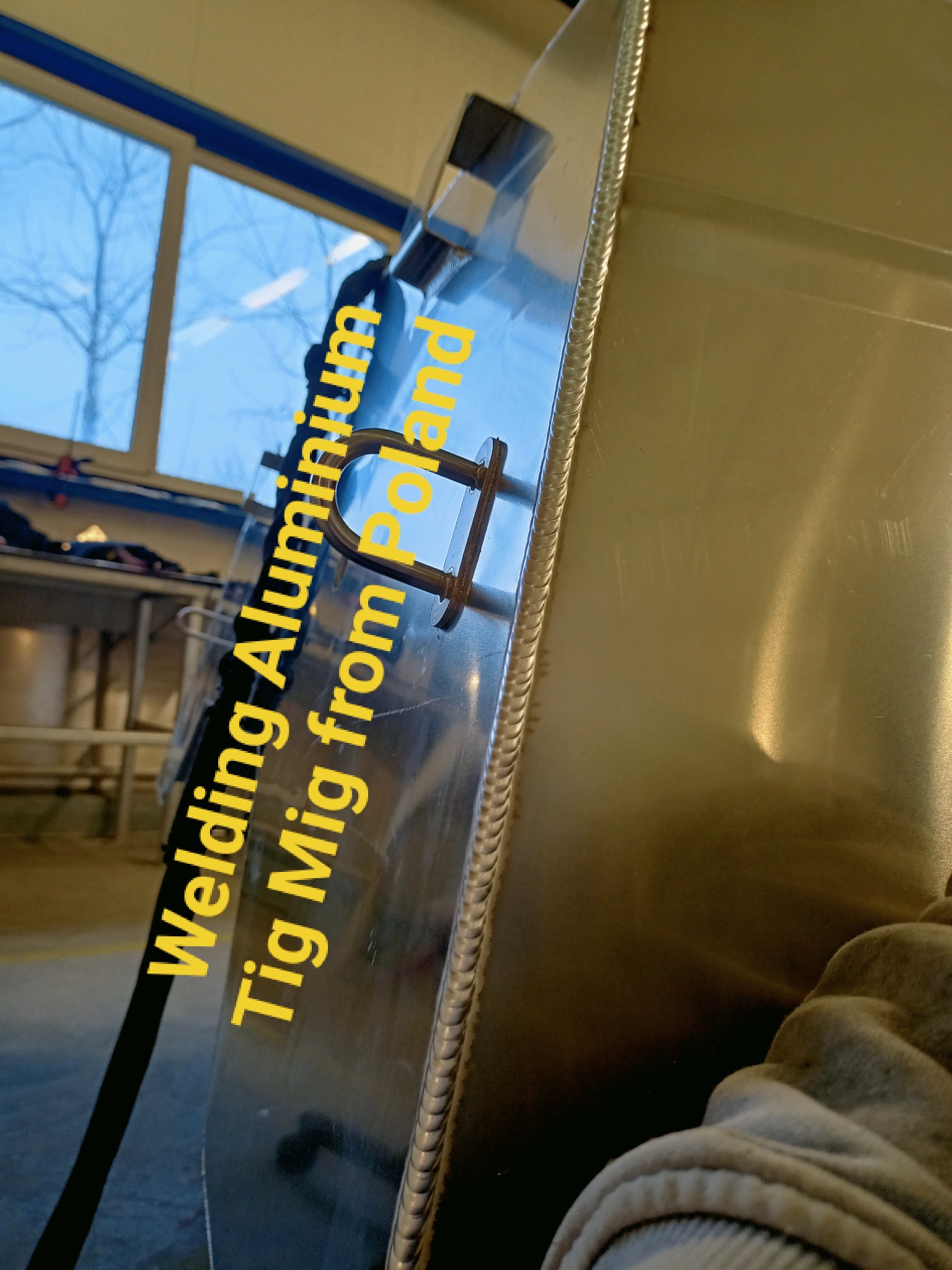 Detal aluminiowej konstrukcji spawanej metodą MIG/TIG, widoczny szew spawalniczy i uchwyt mocujący, w tle okno warsztatu.