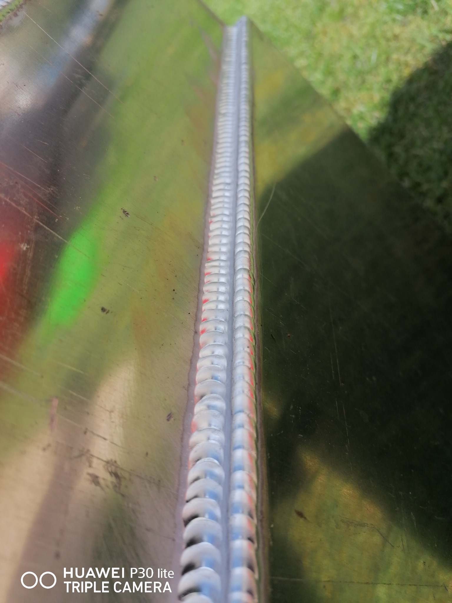 Detaliczny widok spoiny na metalowej powierzchni, widoczne ślady po spawaniu i odbicie otoczenia.