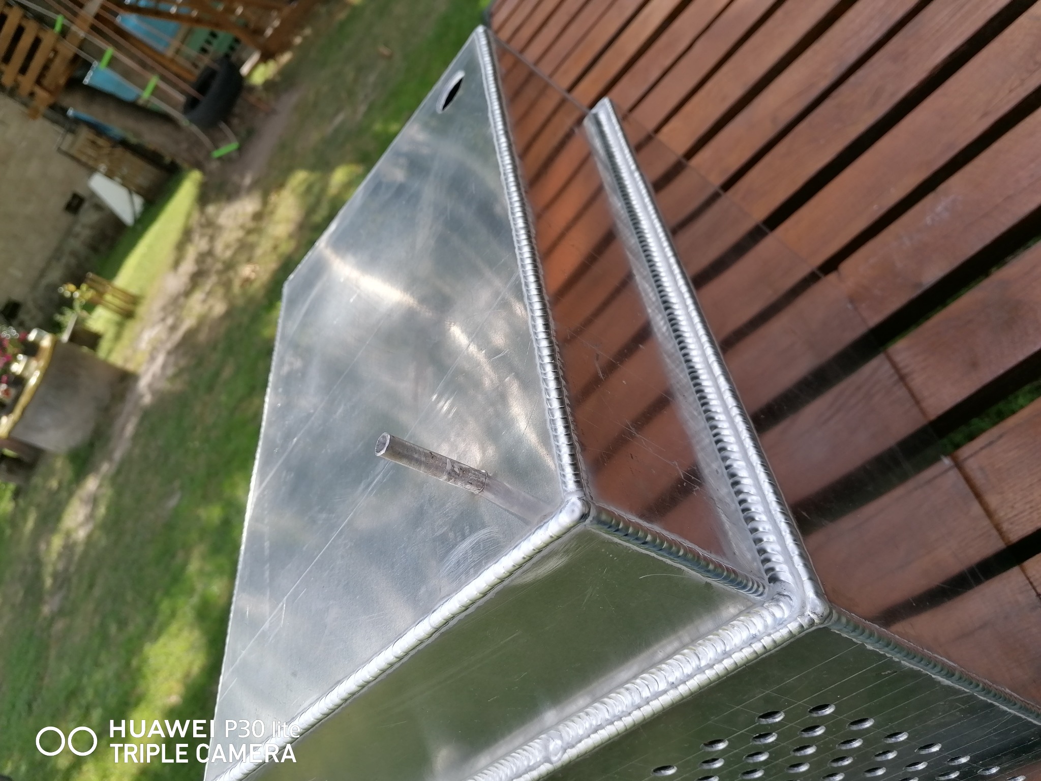 Szczegółowe ujęcie spawów na aluminiowej konstrukcji skrzynkowej z otworami, leżącej na drewnianym podłożu w otoczeniu zieleni.