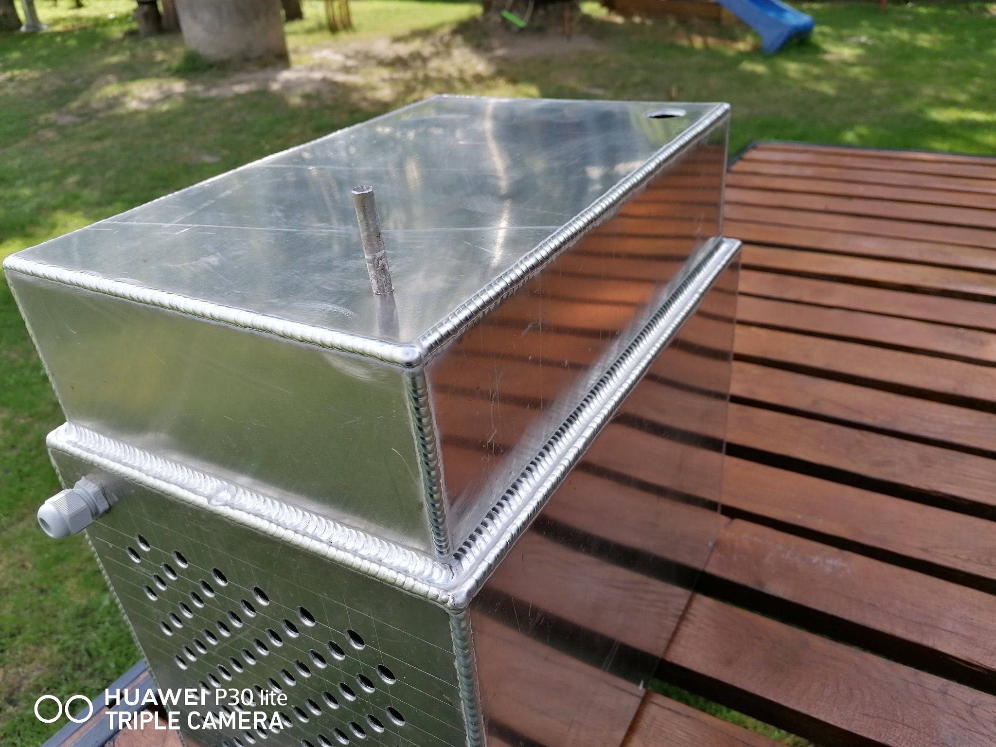 Aluminiowy zbiornik z otworami wentylacyjnymi, spawany metodą TIG, leżący na drewnianym stole na zewnątrz.