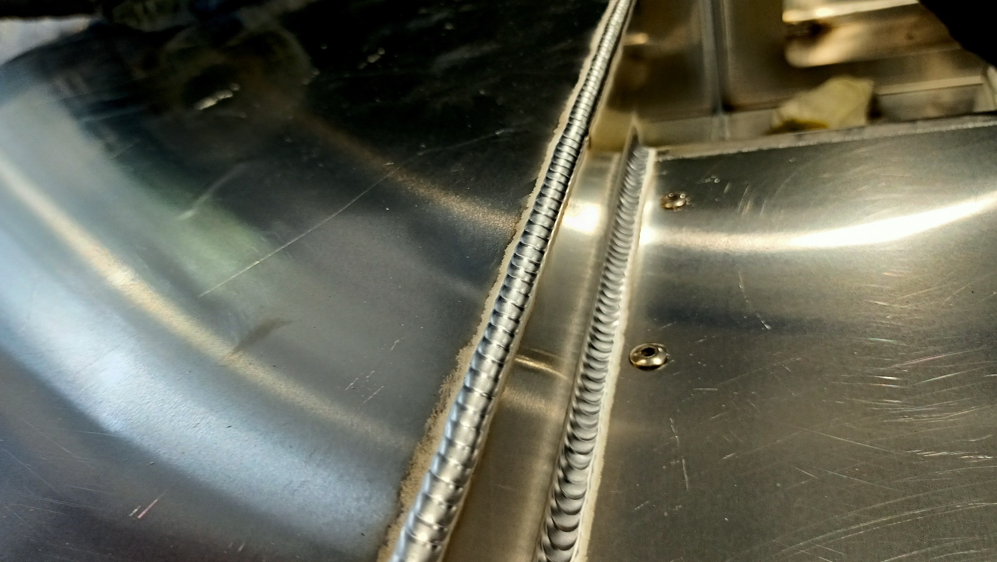 Szczegółowo wykonane spawy na polerowanej, metalowej powierzchni z widocznymi śrubami mocującymi elementy.