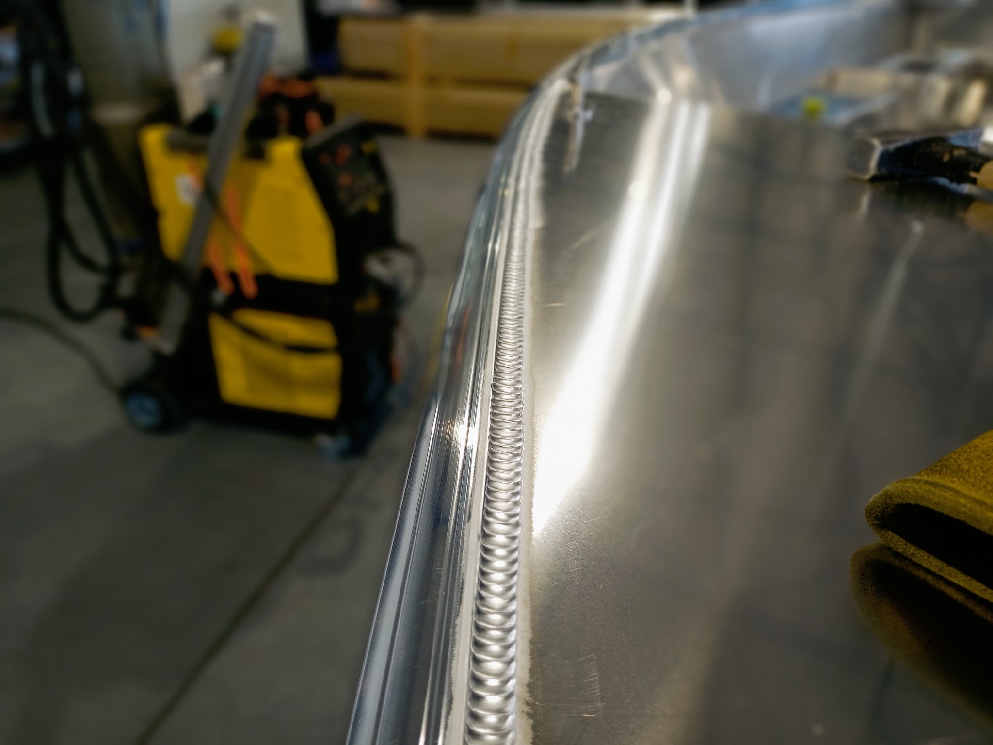 Precyzyjny spaw na polerowanej, aluminiowej powierzchni, widoczny automat spawalniczy w tle.