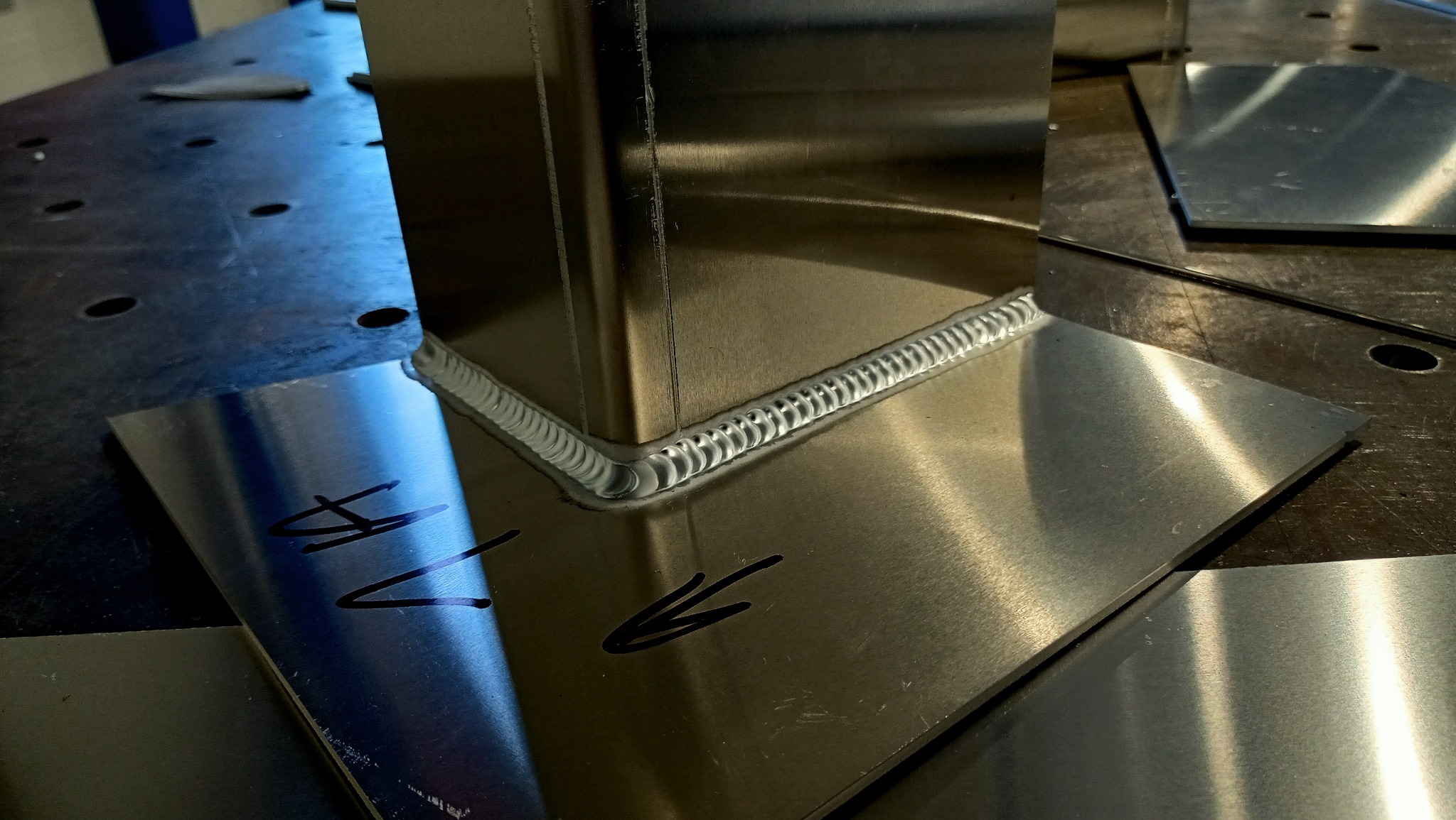 Połączenie spawane dwóch elementów z blachy nierdzewnej pod kątem prostym, widoczny regularny ścieg spawalniczy typu TIG, na jednej z blach widoczne odręczne oznaczenia markerem.