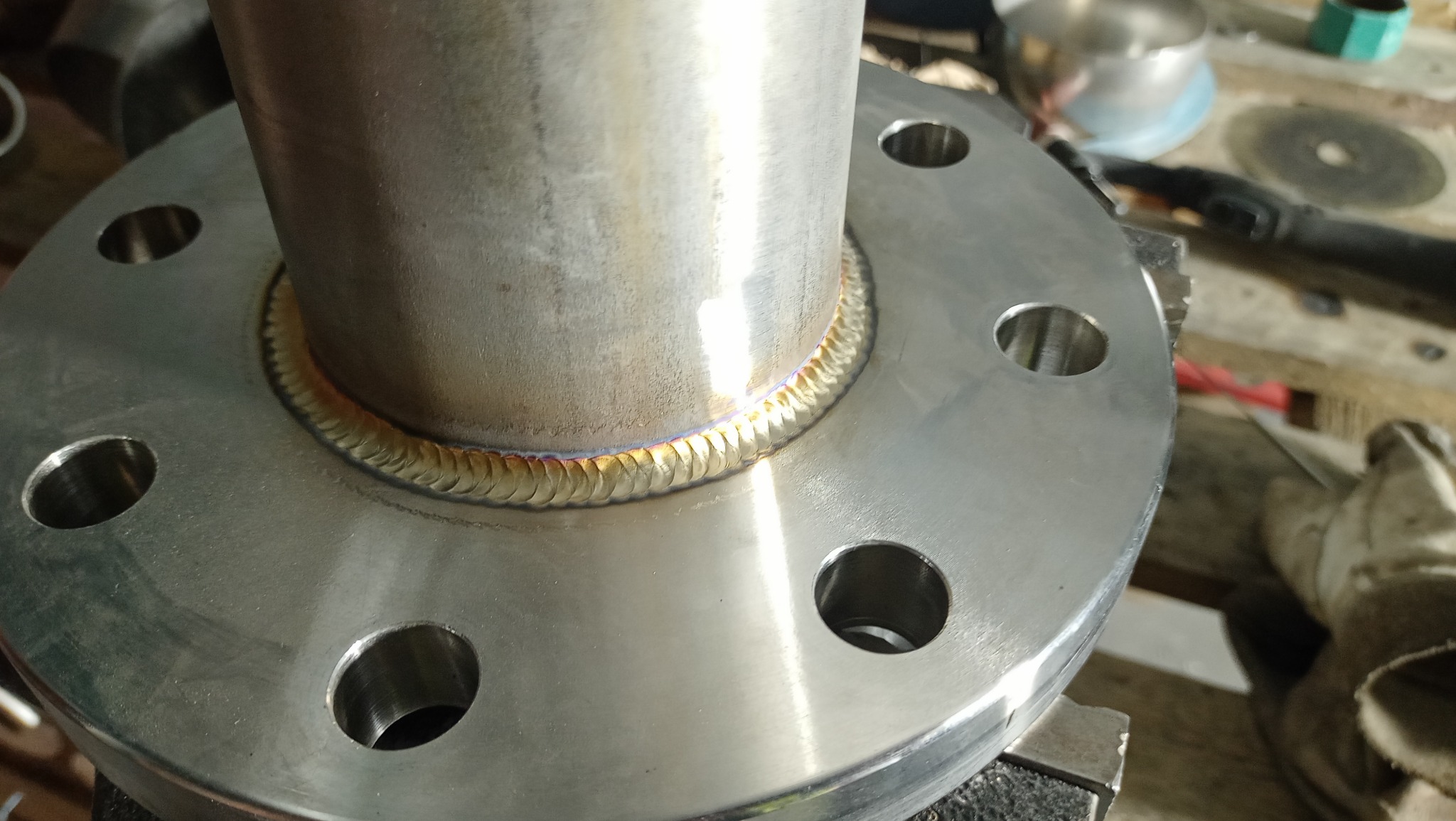 Spoina obwodowa rury ze stalową flanszą, widoczne ślady termiczne i regularny wzór spoiny, tło warsztatowe.