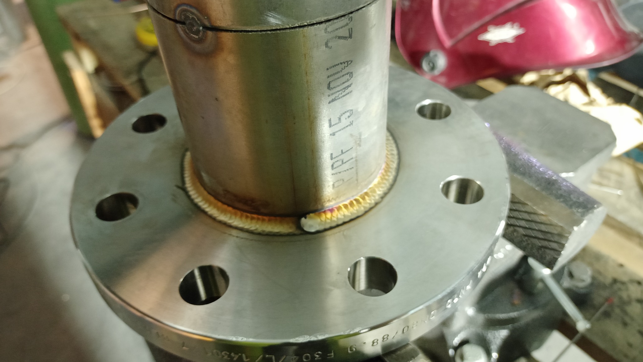 Spoina obwodowa rury ze stali nierdzewnej do kołnierza z widocznymi przebarwieniami termicznymi, z inskrypcją 'PIPE 151 NOV 20' na rurze i oznaczeniami na kołnierzu, uchwycona w uchwycie imadła.