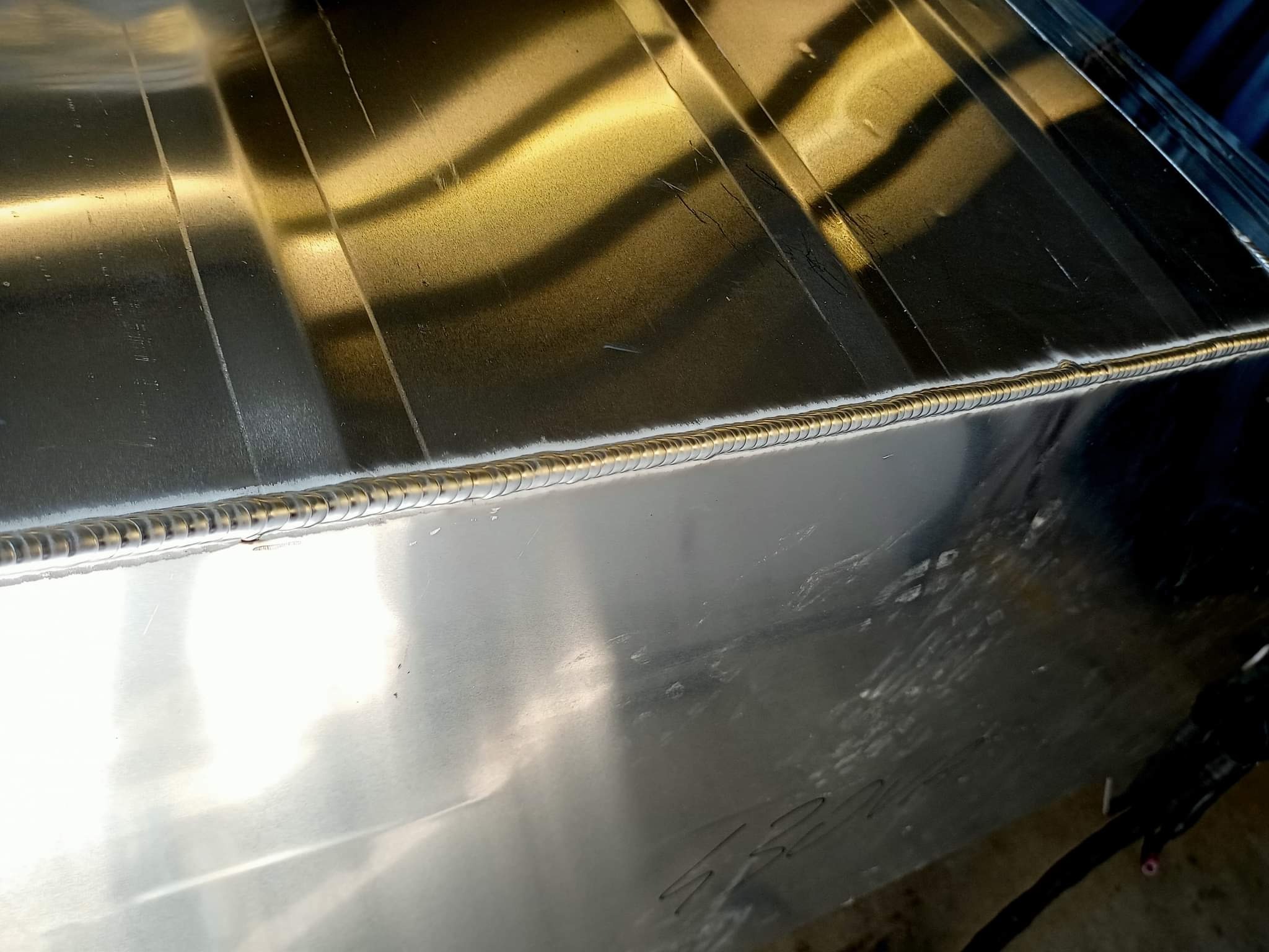 Błyszcząca, srebrna powierzchnia metalowa z widocznym, precyzyjnym spoiwem spawalniczym, tworzącym równy, regularny szew. Widoczne zarysowania i niedoskonałości powierzchni dodają realizmu zdjęciu.