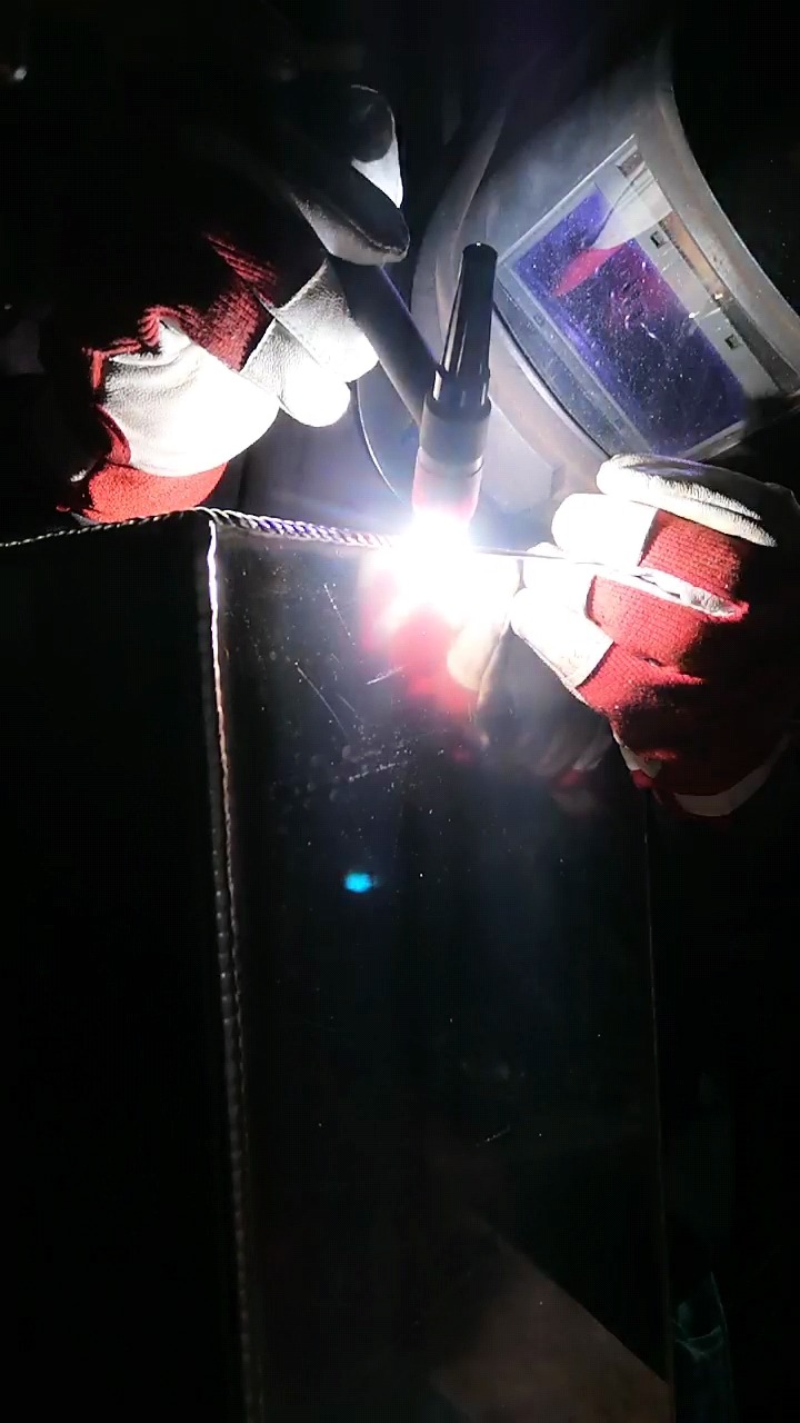 Zbliżenie na proces łączenia metalu metodą TIG, widoczne iskry i czerwone rękawice ochronne na dłoniach operatora, spawanie narożnika metalowej konstrukcji.