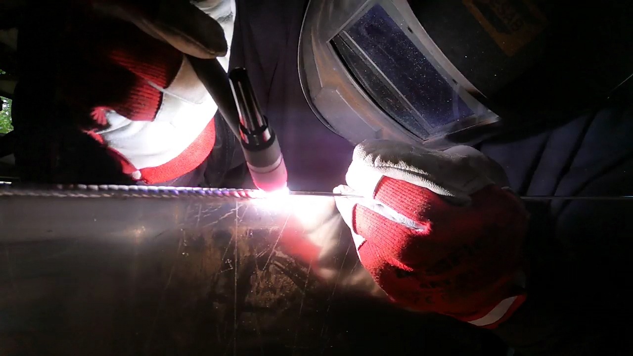 Zbliżenie na proces łączenia metalu metodą TIG, widoczny jasny łuk elektryczny, elektroda wolframowa i rękawice ochronne z czerwonymi detalami.