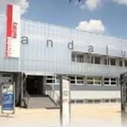 Fasada budynku usługowego z szarej blachy trapezowej, z dużym czerwonym panelem reklamowym z nazwą 'Andaluzja', schody prowadzące do wejścia, plakaty reklamowe przed budynkiem, błękitne niebo w tle.