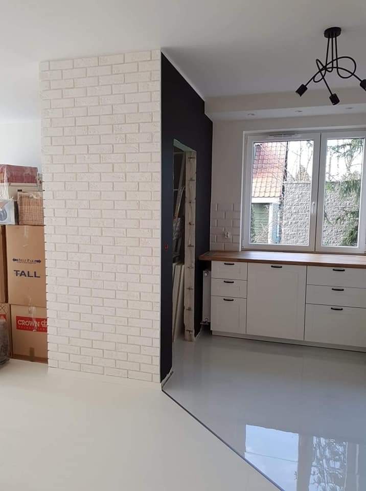 Biała cegła dekoracyjna na ścianie w nowocześnie wykończonym wnętrzu, widoczne kartony z rzeczami, białe szafki kuchenne z drewnianym blatem i czarna ściana obok drzwi.