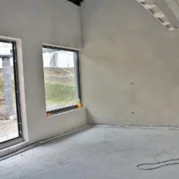 Wnętrze surowego stanu z otynkowanymi ścianami, widoczne dwa okna z czarnymi ramami, betonowa podłoga i sufit, butelka z płynem w niszy okiennej.