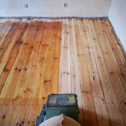 Częściowo wycyklinowana drewniana podłoga z widoczną maszyną do cyklinowania i workiem na pył, ukazująca różnicę w kolorze i strukturze drewna przed i po obróbce.