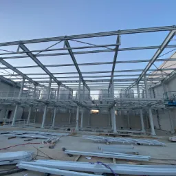 Szkielet hali produkcyjnej w trakcie budowy, widoczne stalowe elementy konstrukcyjne, zbiorniki, podnośnik koszowy i materiały budowlane na placu budowy.