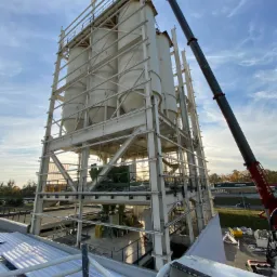 Wysoka, biała konstrukcja wsporcza z trzema cylindrycznymi zbiornikami montowana za pomocą dźwigu teleskopowego na tle błękitnego nieba z chmurami.